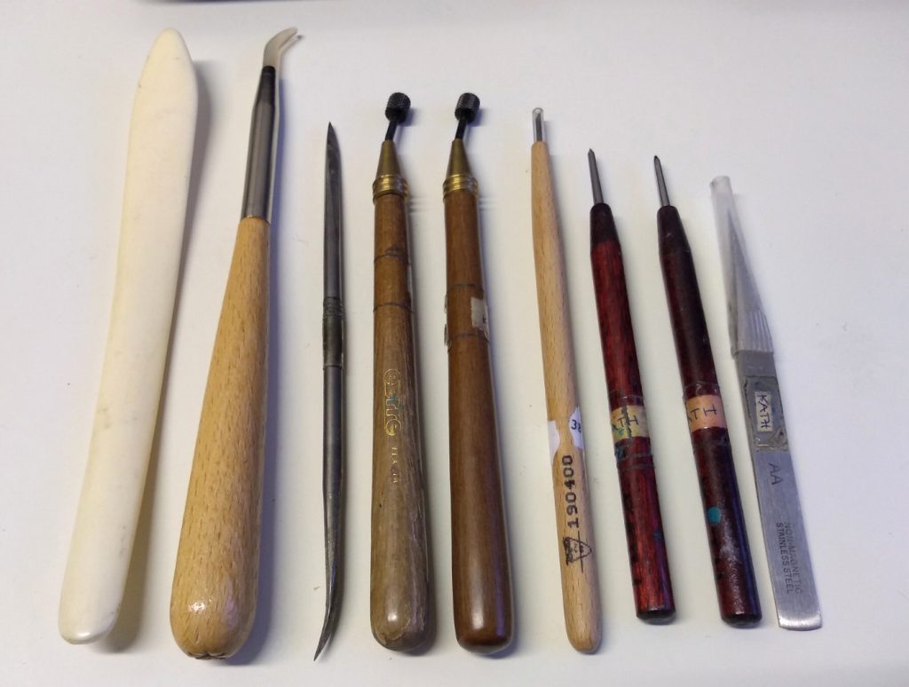 Kath's etching and printing tools which include from left to right: a bone folder, agate burnisher, steel scraper and burnisher, two roulettes with different sized dot patterns, square tipped etching needle, sharp pointed etching needle, rounded point etching needle and a pair of jeweller's tweezers. 
