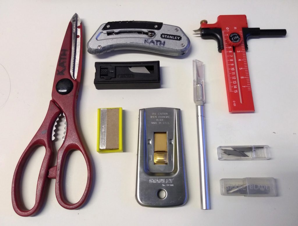 Kath's sharps collection: Scissors, cutting knife, scalpel, paint scraper, and compass cutter. There are also spare blades for each cutting tool. 